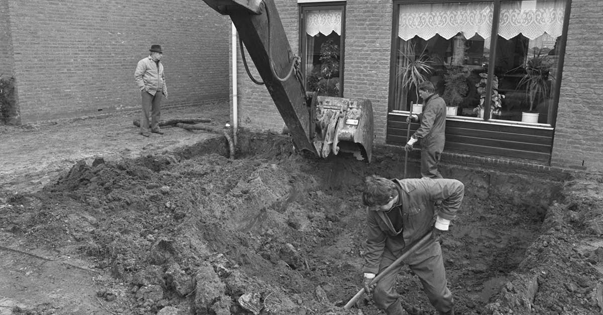 Zwart-wit foto van een afgraving van een voortuin, voor het huis is een kuil te zien met daarin twee mannen en een graafmachine die de grond afgraven.